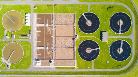 Vue aérienne d'une station d'épuration avec des bassins circulaires noirs et rectangulaires marron, entourée de pelouses vertes.