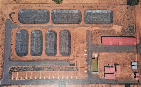 Vue aérienne de bassins aquacoles recouverts de bâches noires à côté de bâtiments rouges sur un terrain sec.